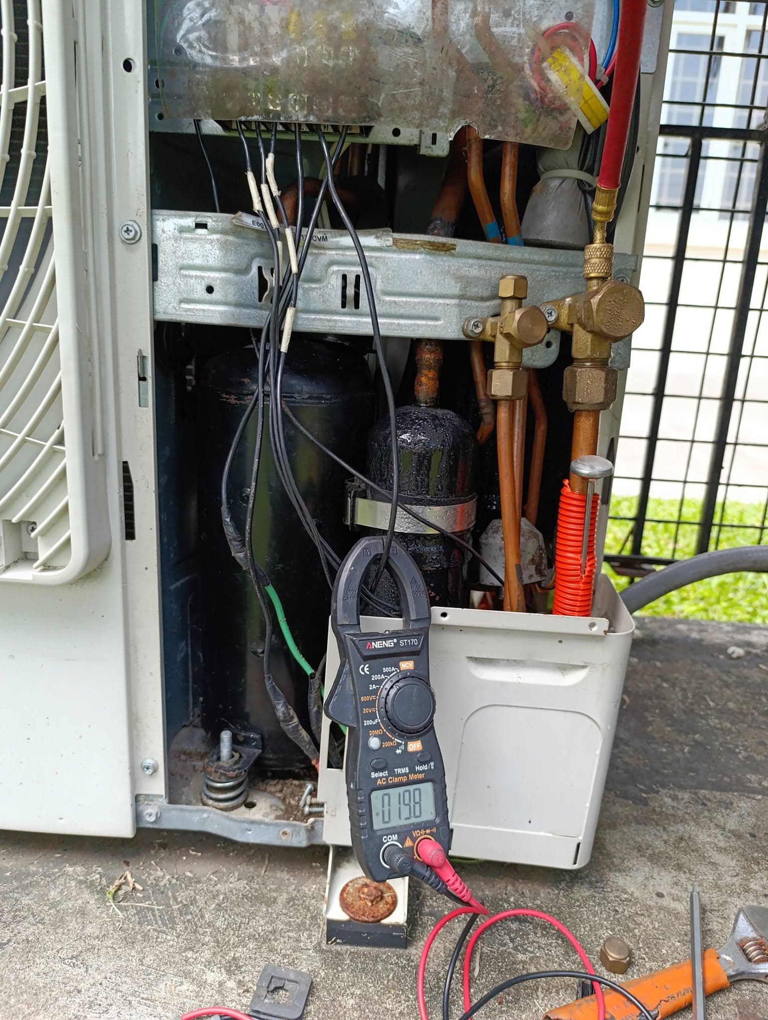 Open inverter air-conditioning unit undergoing electrical repair testing with a clamp meter.